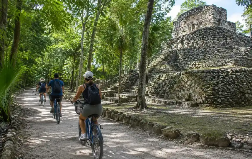 Visitors riding bicycles on a shaded jungle path beside a Coba stone temple
