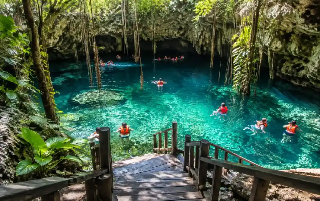 Swimmers in life vests below wooden stairs in a clear turquoise jungle cenote